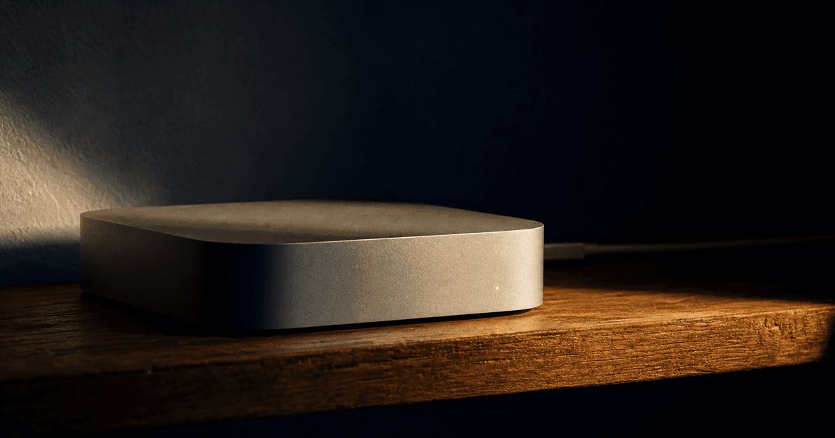 A small aluminum desktop computer on a shelf, single amber status light glowing, network cable fading into shadow